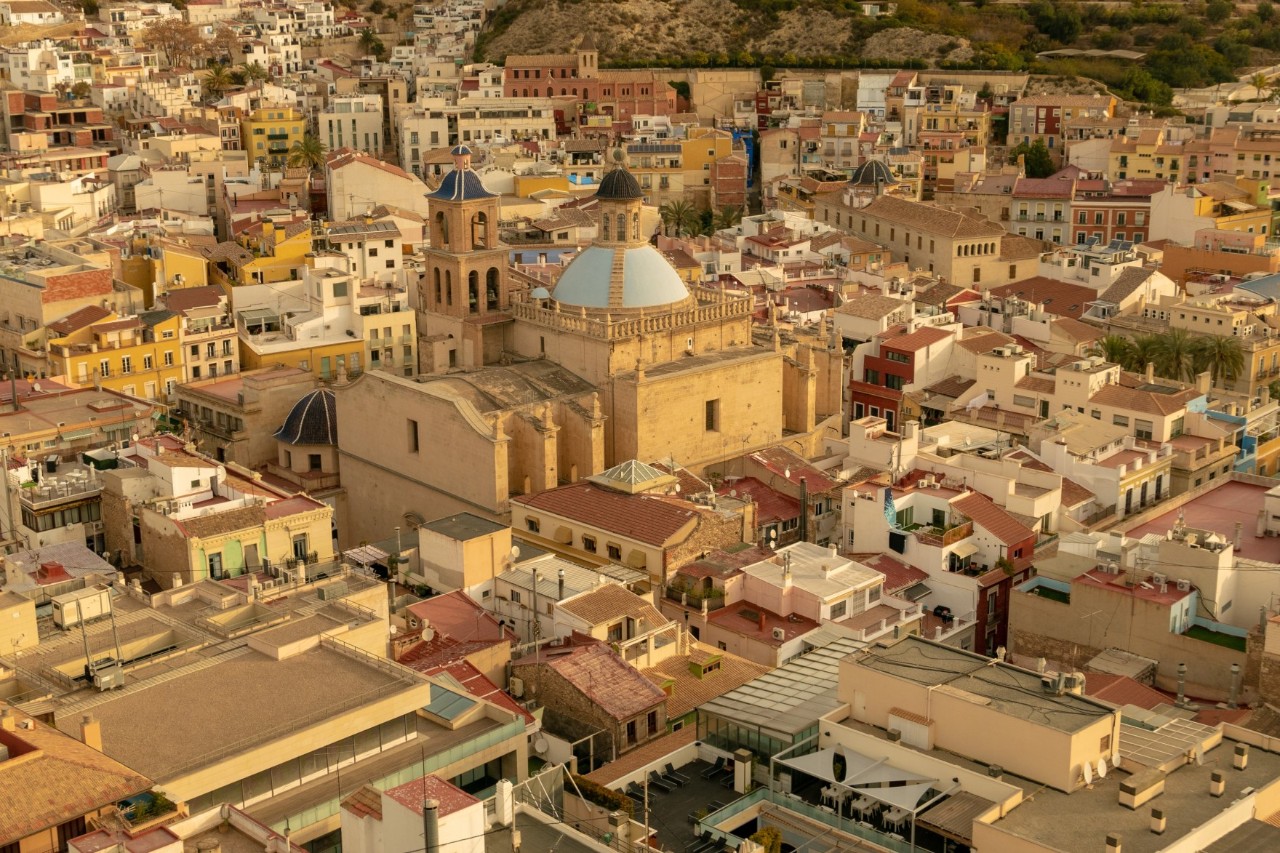 Aerial view of a densely built-up old town, glowing warmly in yellowish colours in the sunlight. In the centre of the picture is an ochre-coloured cathedral with a larger light blue and a smaller dark blue dome.