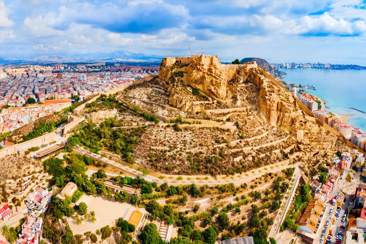 Aerial view of a fortress built on a mountain above a city. A winding path leads up the mountainside. The city can be seen below the fortress and the sea on the right of the picture.