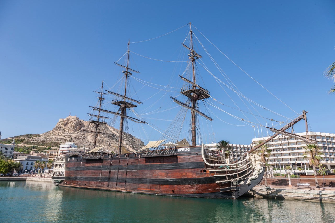 View of a historic sailing ship moored in a harbour. The ship is made of wooden planks and has three masts, a bowsprit and a figurehead. The sails are retracted. Modern buildings and a mountain can be seen in the background.