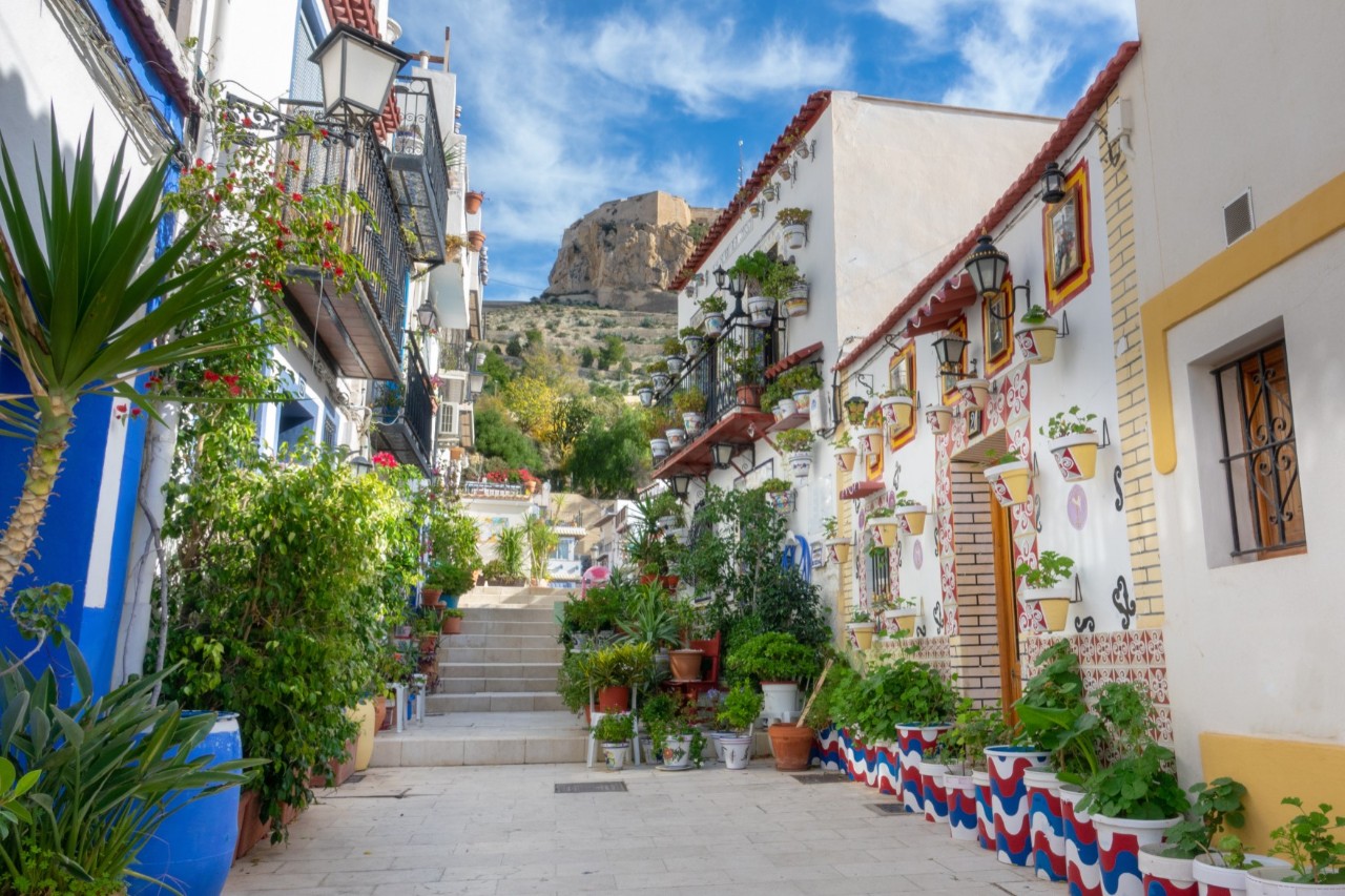 View of an old-town street with steps leading upwards. The white buildings on both sides of the street are colourfully decorated with mosaic elements and bright, colourful accents. The walls of the buildings and the street are decorated with numerous plant tubs, some of which are also colourful.