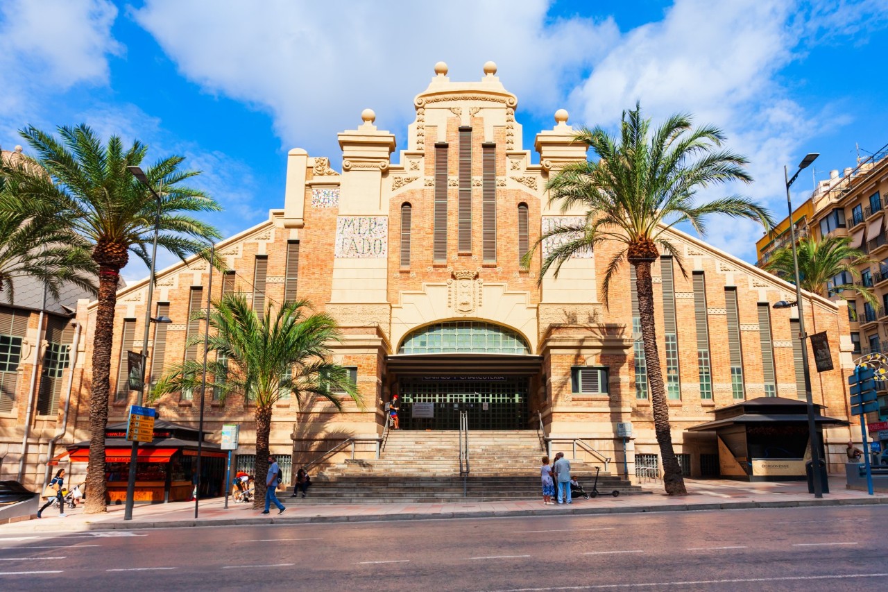 Terracotta-coloured front of a large building, reminiscent of a church front with sloping wings and high windows. The entrance consists of a large gate. There are palm trees directly in front of the building and a square in front of it.