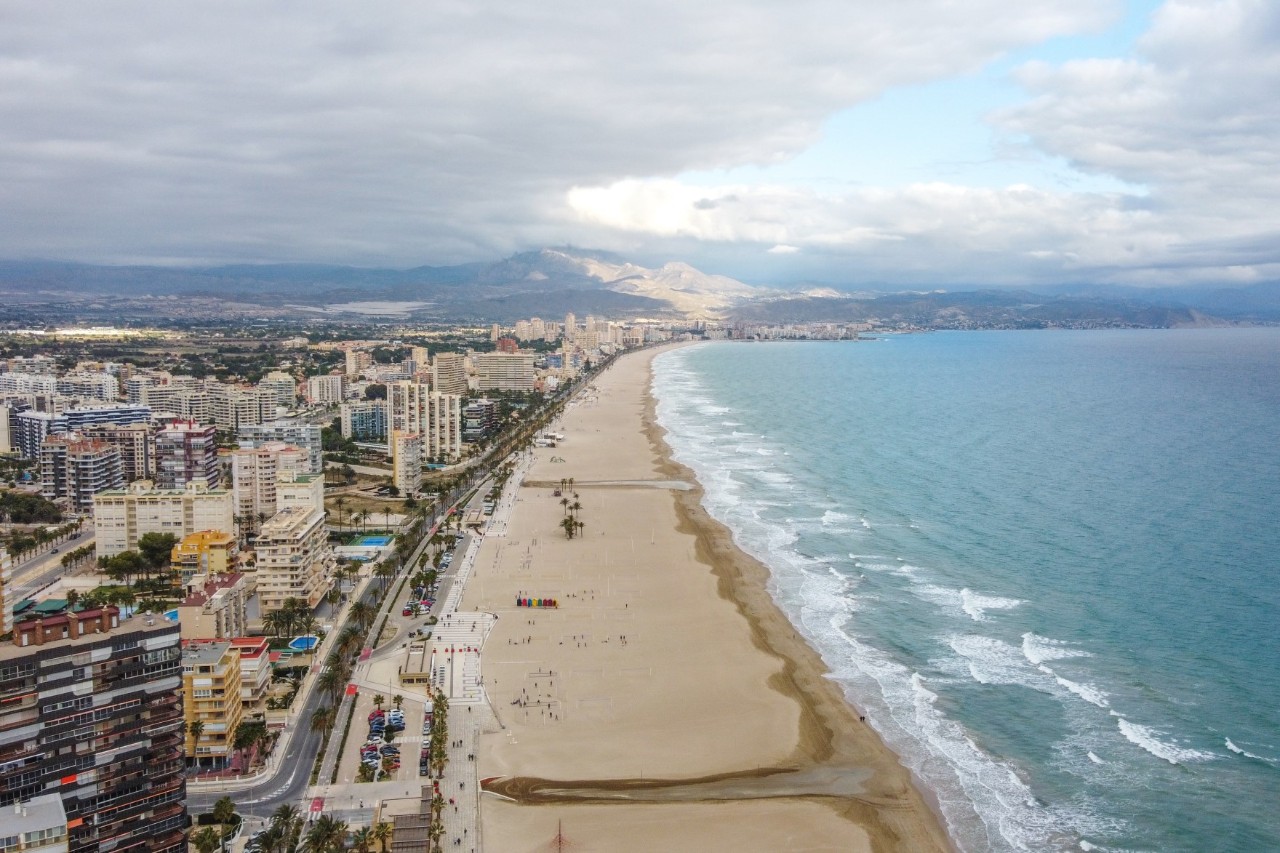 Aerial view of a long beach directly on the outskirts of the city with light sand and turquoise water. On the left of the picture part of a larger city with numerous high-rise buildings can be seen along the beach.