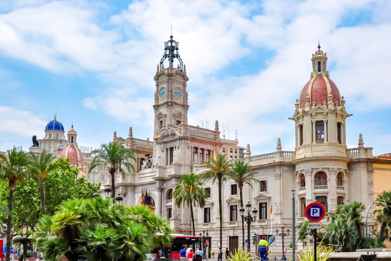 Side-front view of a historic, light-coloured building with a decorated front, a tower with a clock in the centre and towers with red domes on either side. There are palm trees in front of the building. A few people and a bus can be seen in the foreground.