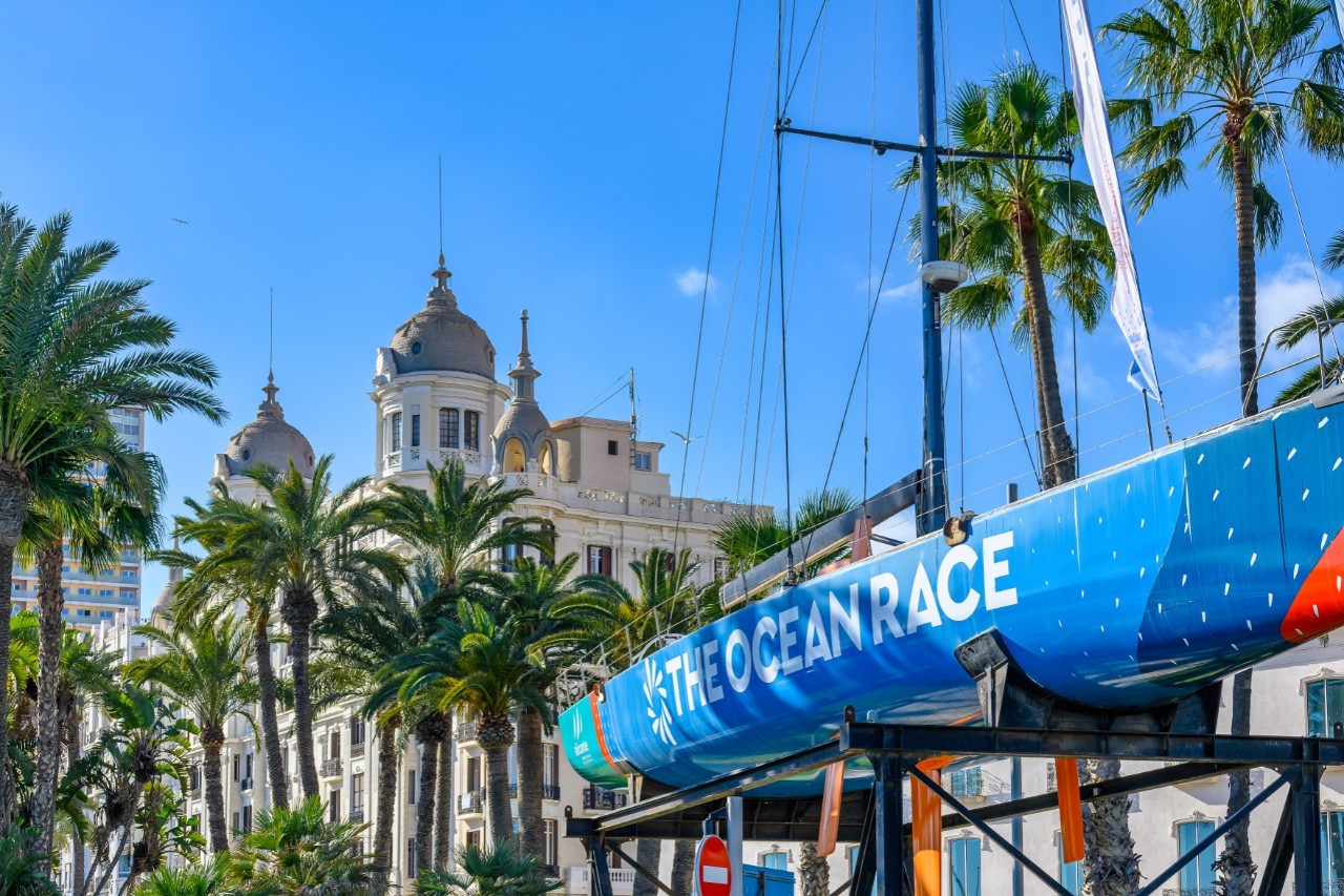 A blue sail boat with retracted sails, lying on a rack on land with “The Ocean Race” written on it in large letters. In the background is a striking white building with a domed tower and palm trees in front of it.