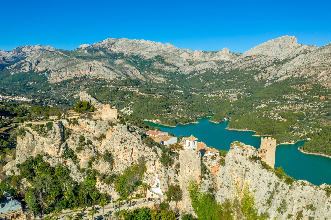 Aerial view of a mountain village with a few buildings and an old tower situated on a mountain range. Below is a turquoise reservoir and another mountain range on the horizon.