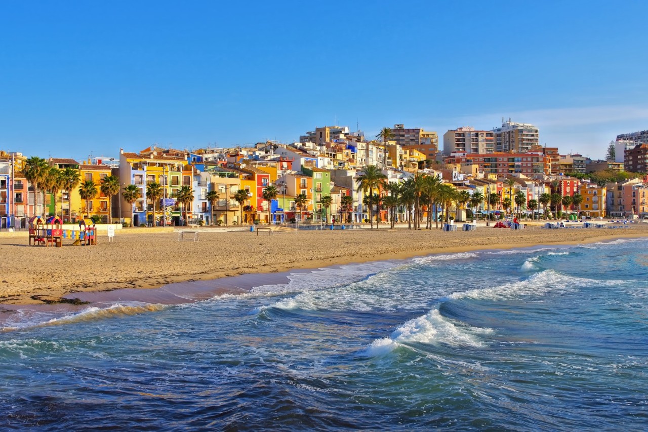 View from the sea of a colourful row of buildings directly on a sandy beach with a few high-rise buildings visible in the background. The promenade in front of the row of buildings is lined with palm trees.