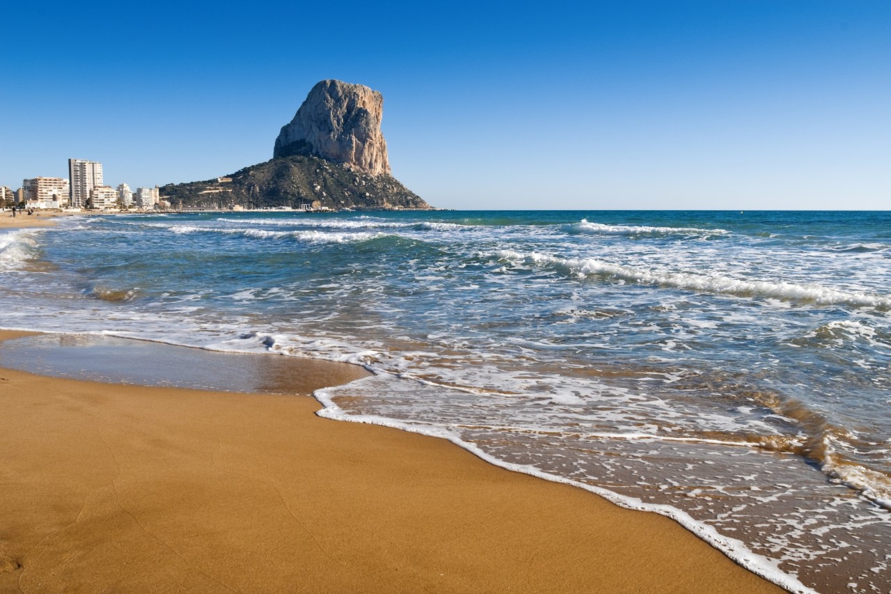 View from a sandy beach with wavy sea to a huge rock with a flattened top in the background. At the foot of the rock you can see the town’s buildings.