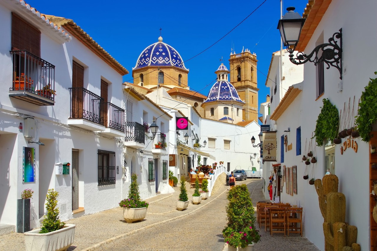 View into a small street in an old town with white, two-storey buildings on both sides. There are several plant pots in the street. The blue domes of a church and a tall tower can be seen in the background.