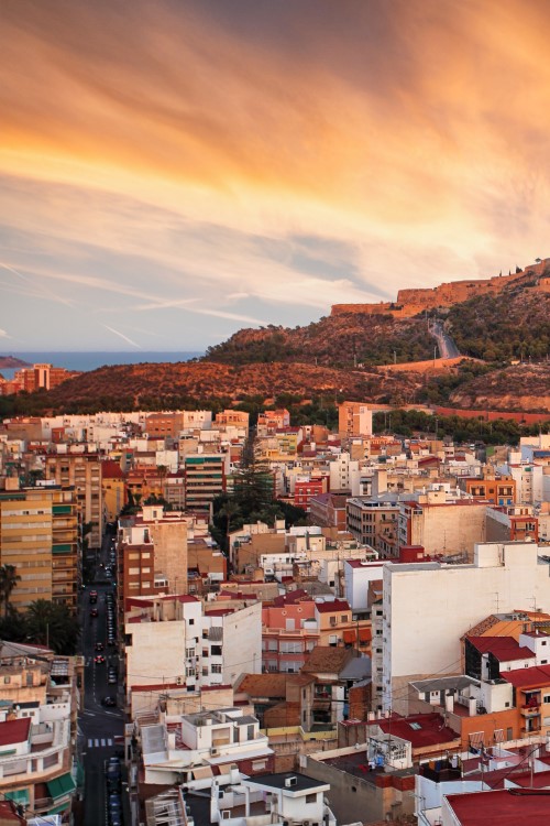 Aerial view of Alicante at sunset. In the foreground is a sea of buildings illuminated red by the sunset, in the background a mountain with a fortress on the summit, which also glows red.