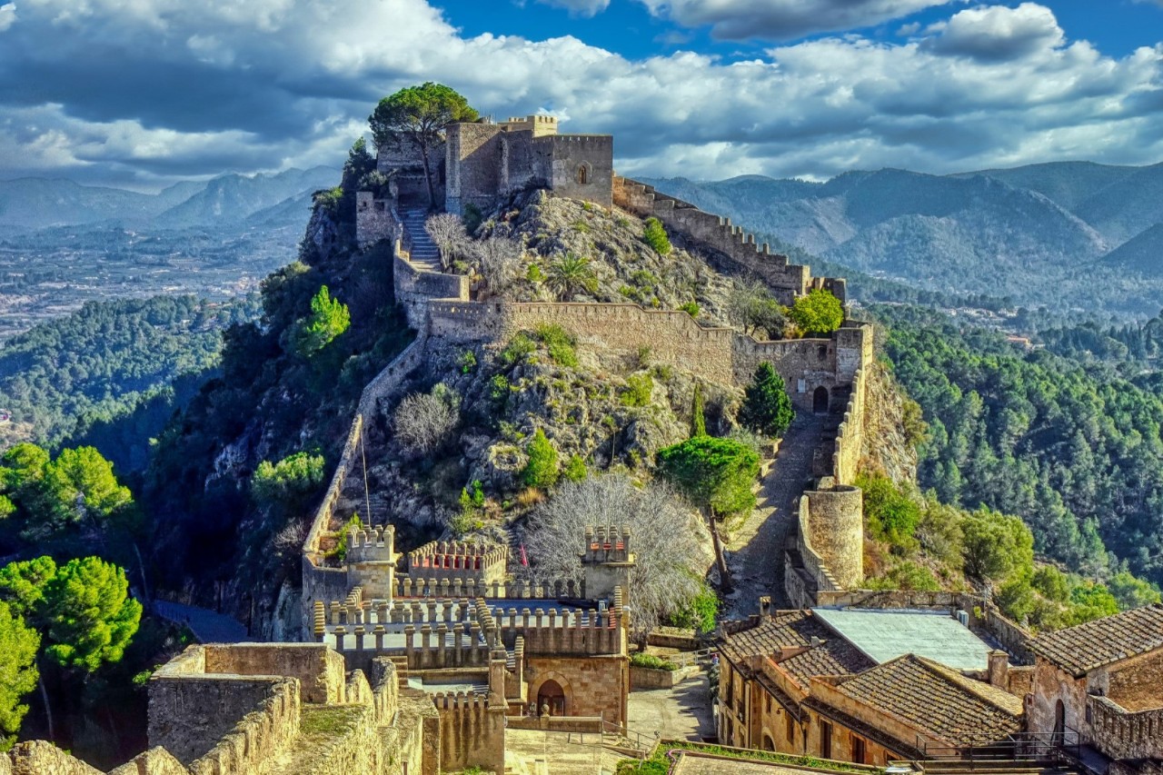 Imposing castle complex that extends over a rocky hill. The castle walls are lined with towers. Wooded hills and valleys can be seen in the background.