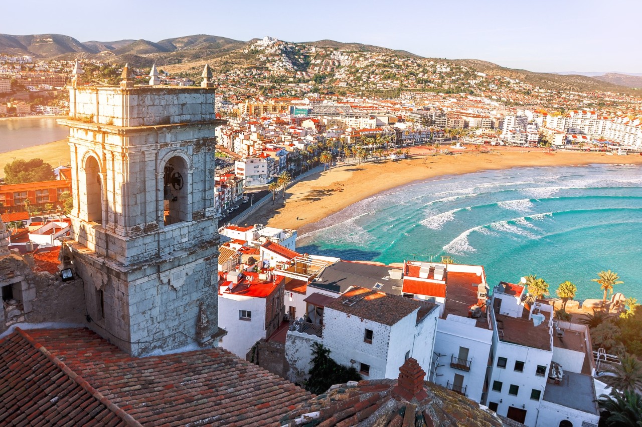 The picture shows a coastal town with a curved sandy beach from a bird's eye view. The town consists of many white houses. Hills with more houses can be seen in the background. The sea has clear, turquoise-coloured waves.