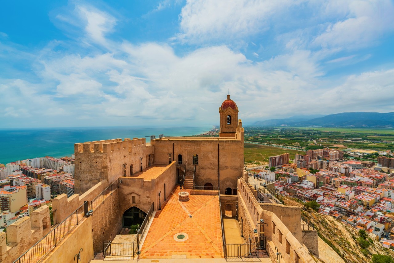 The picture shows a beige-coloured, medieval castle with a large terrace surrounded by a wall and a small bell tower. A wide coastal landscape with the sea and a village can be seen in the background.