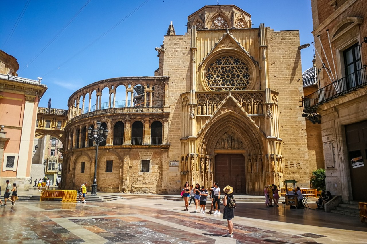 The picture shows the façade of a cathedral with a large rose window above the entrance. The round arches of a neighbouring building can be seen next to it. In front of the cathedral, there are people on a square with a marble floor. 
