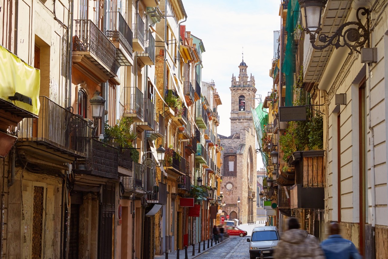 The picture shows a narrow street lined with multi-storey older buildings with balconies. Some of the balconies are decorated with plants. A historic church tower can be seen in the background. 