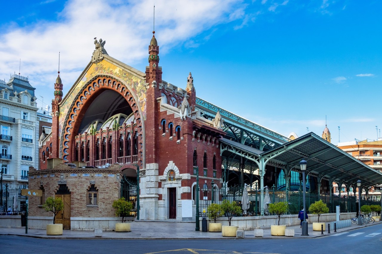 The picture shows a historic market hall building with red bricks, ornate towers and a large archway with mosaic decoration. Modern buildings can be seen on the left and a steel and glass construction of a market hall on the right.