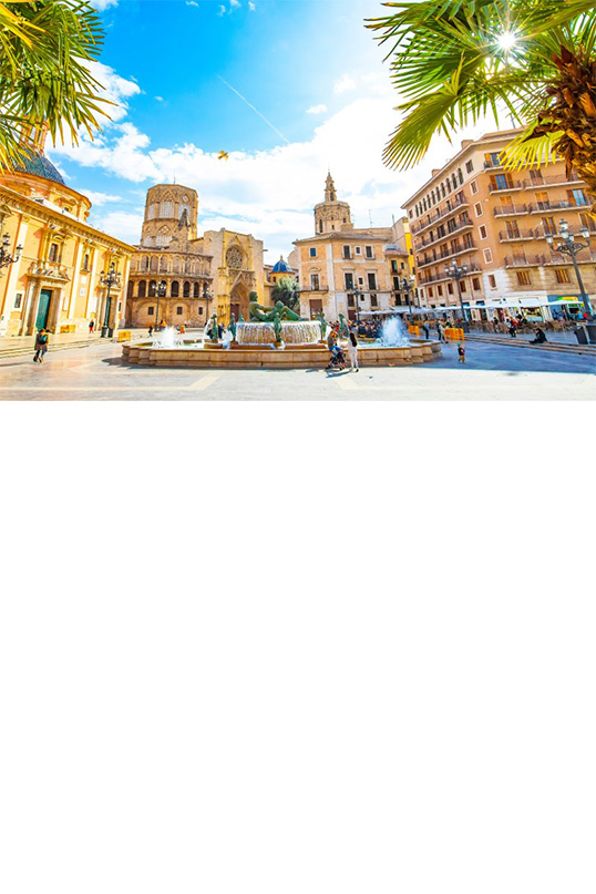 The picture shows a sunny, lively square with palm trees, a Gothic cathedral, a striking tower and other historic buildings. In the centre is a fountain with a large statue surrounded by bubbling water. 