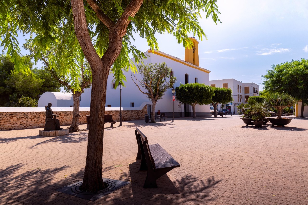 The picture shows a sunny square with a paved floor, surrounded by trees and plants. In the background, there is a white church with a yellow bell tower. A bench and a statue are in the shade of the trees.