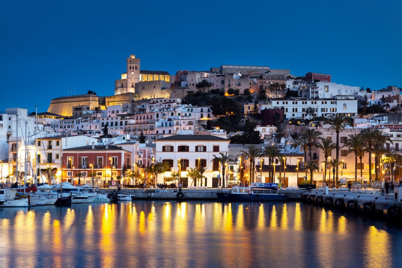 The picture shows a city on a hill at night. In the foreground, there is an illuminated harbour with boats. The city walls and the cathedral tower over the whitewashed houses, and lights reflect in the water.