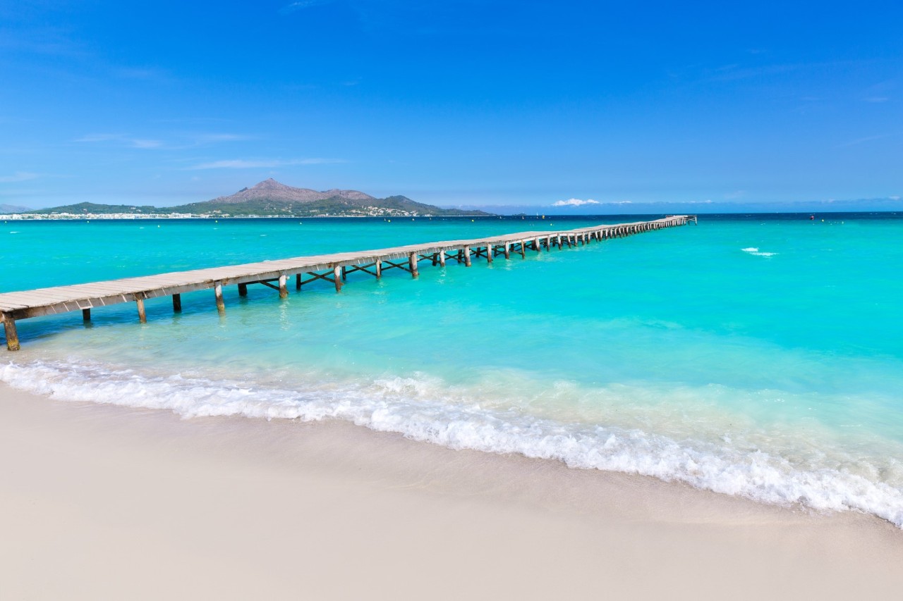 Turquoise water and a white sandy beach with a long jetty leading into the water and a mountain view in the background © lunamarina/stock.adobe.com
