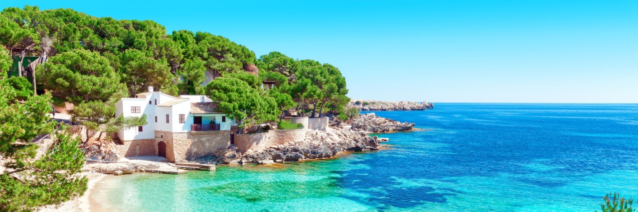 Turquoise water and a white sandy beach on Majorca, surrounded by trees and a white house © pixelliebe/stock.adobe.com