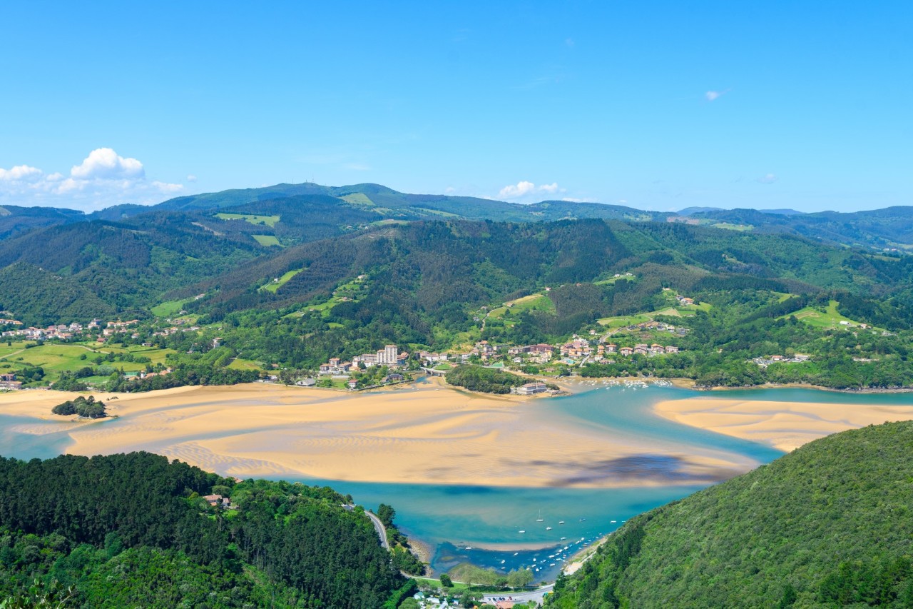 The picture shows a river estuary with sandy banks and turquoise blue water. The landscape is surrounded by green hills, forests and scattered houses.