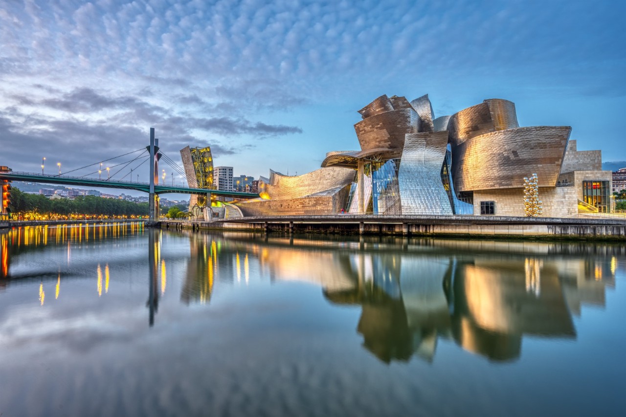The picture shows an imposing building with curved shapes made of titanium, glass and limestone. It lies on the riverbank and is reflected in the water. A modern bridge can be seen on the left of the picture.