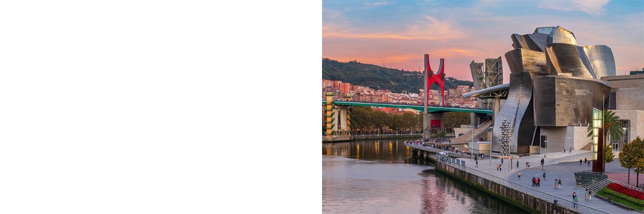 The Guggenheim Museum in Bilbao with its curved architecture made of titanium and glass, surrounded by a modern bridge and a promenade along the river with walkers and palm trees. 
