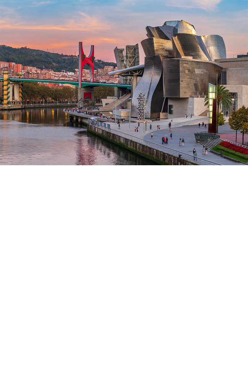 The Guggenheim Museum in Bilbao with its curved architecture made of titanium and glass, surrounded by a modern bridge and a promenade along the river with walkers and palm trees. 