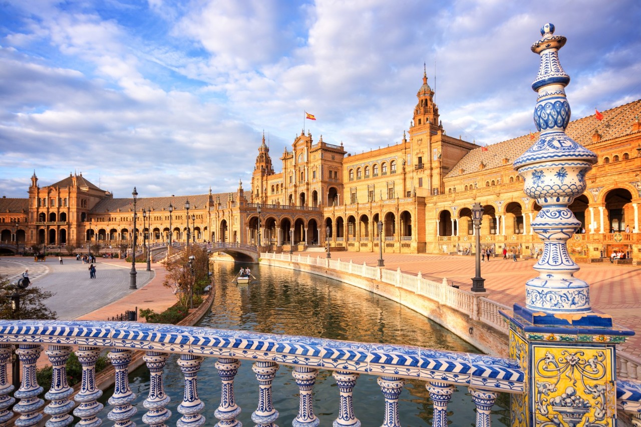 The picture shows an impressive semicircular building with arcades, towers and a canal. In the foreground, you can see elaborately decorated blue and white ceramic railings. 
