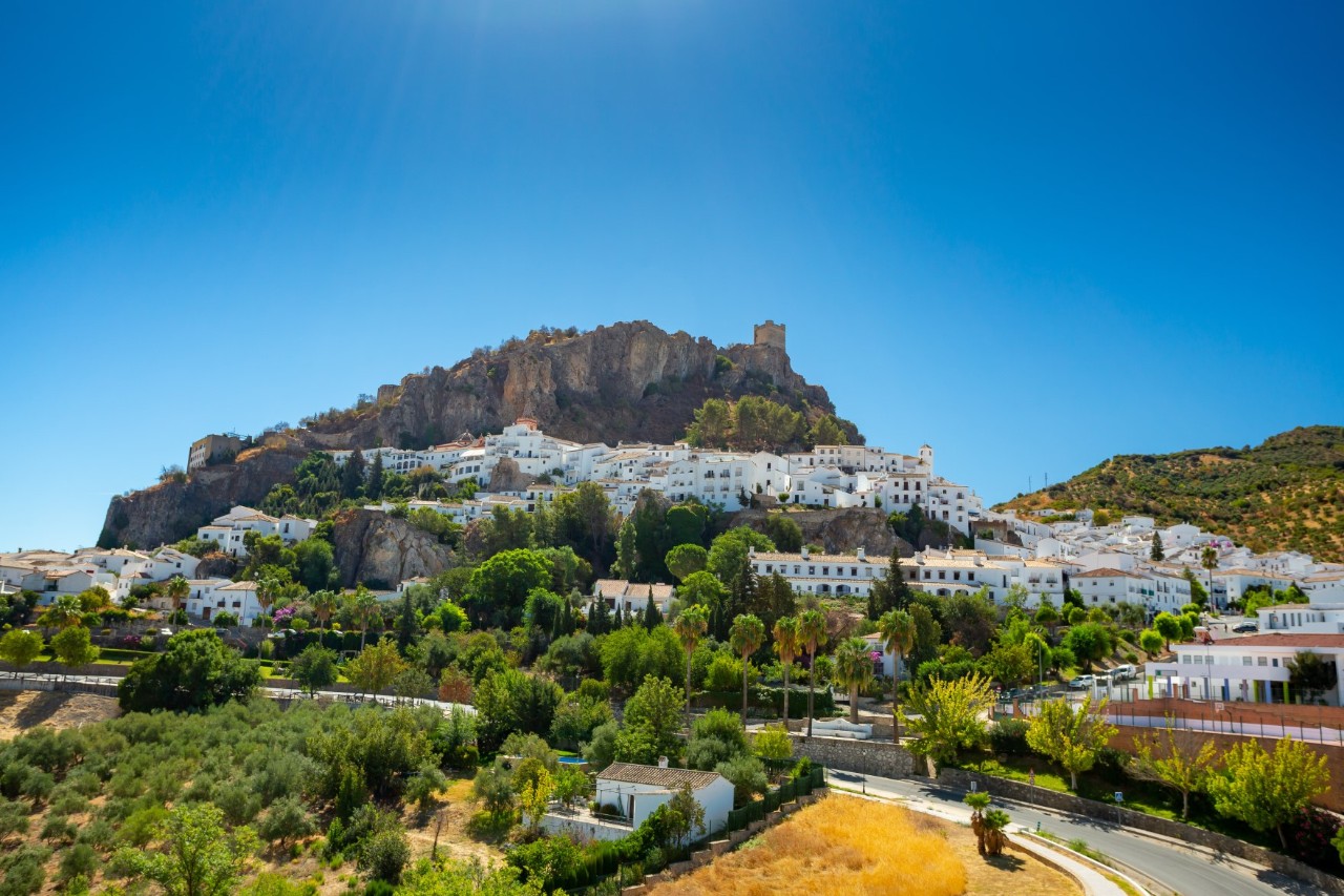The picture shows a village on a mountainside with a castle at its summit. The white houses blend harmoniously into the rocky landscape, surrounded by greenery and Mediterranean plants.