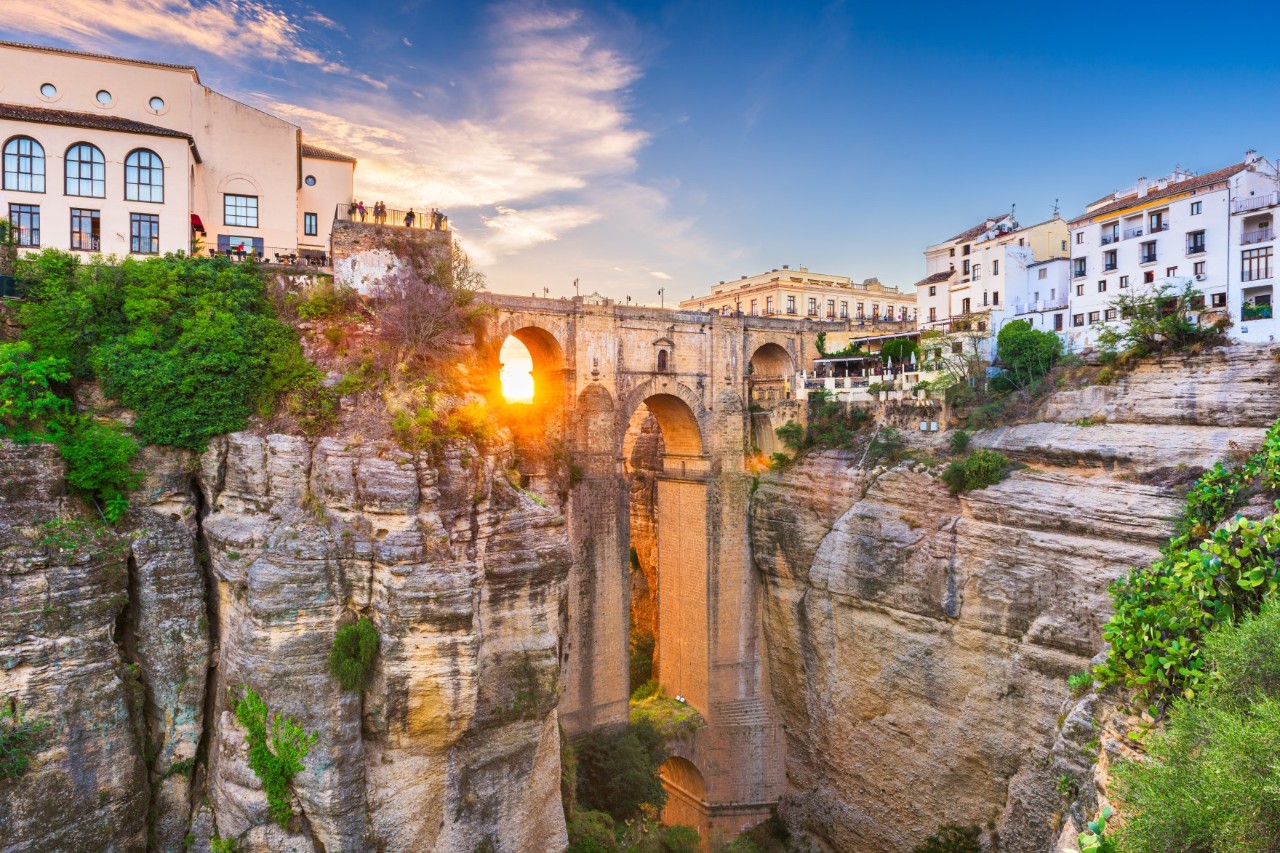 The picture shows an impressive stone bridge spanning a deep gorge. The sun shines through one of the arches, while white houses and green vegetation line the rocky cliffs.