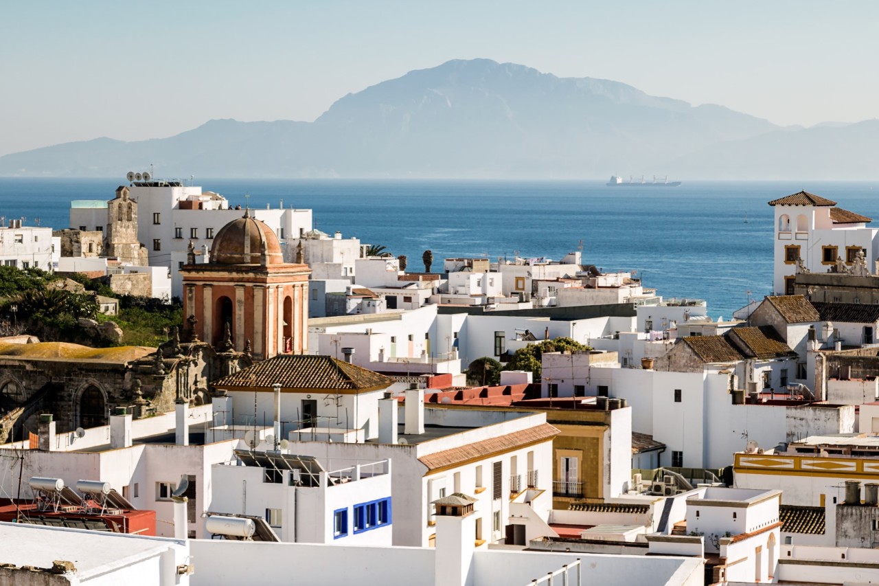 The picture shows a coastal town with white houses and a historic church in the foreground. In the background, the deep blue sea extends to a coast and a passing ship.