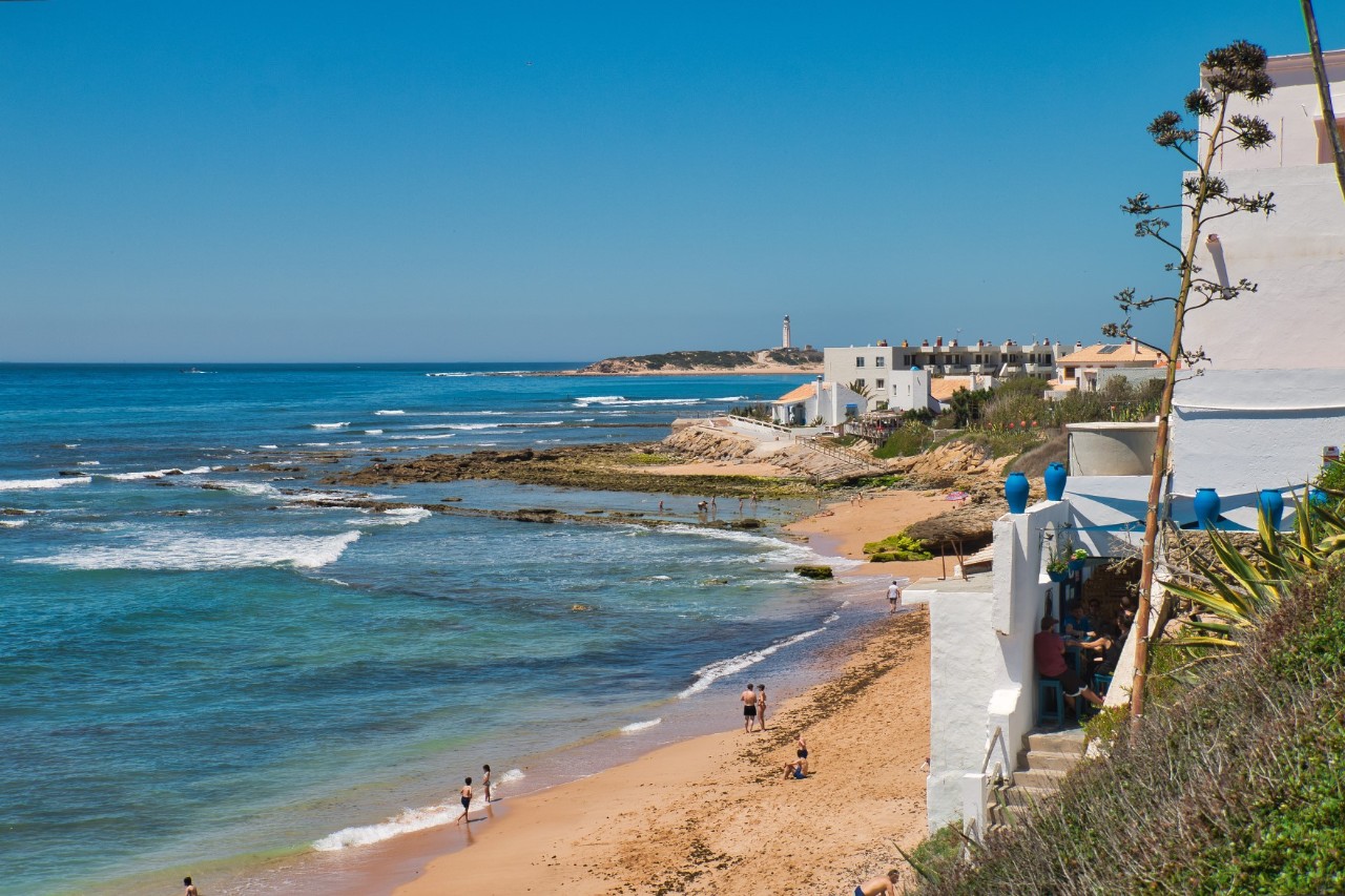 The picture shows a coastal landscape with a blue sea and a narrow sandy beach with a few people. Scattered white houses with blue accents can be seen on the right. 