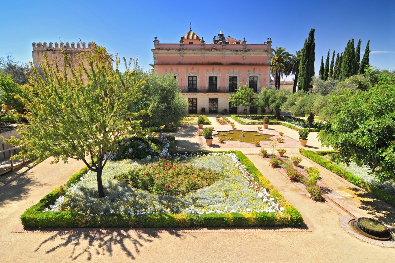 The picture shows a magnificent garden with flowerbeds, well-kept sand-coloured paths and a small fountain. In the background stands a building with a light pink façade. Cypress trees and a medieval fortress wall complete the picture.
