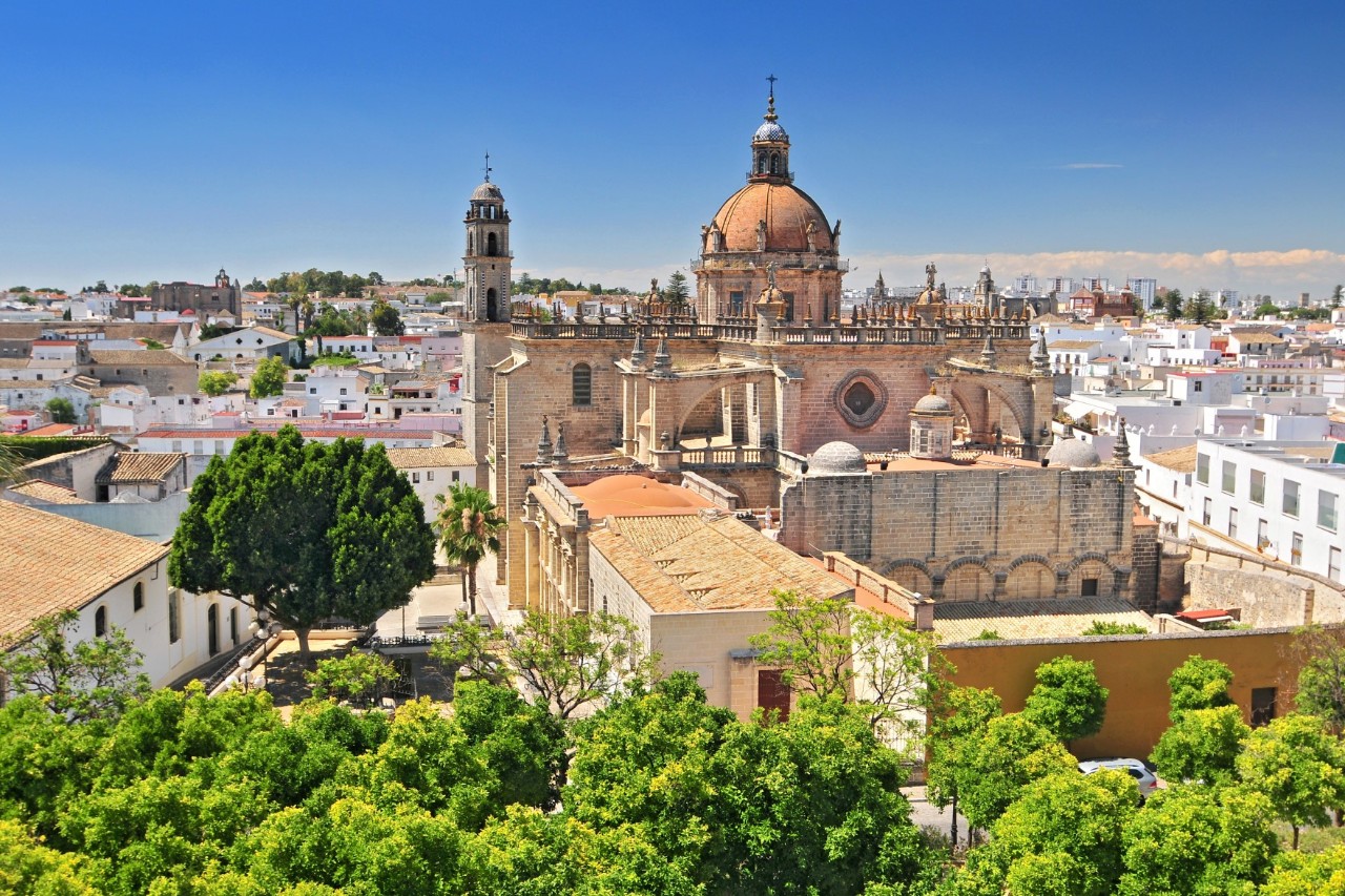 The image shows a massive cathedral with a large red dome, towers and adjoining church buildings, embedded in a Mediterranean urban landscape. In the foreground, there are green trees and white houses.