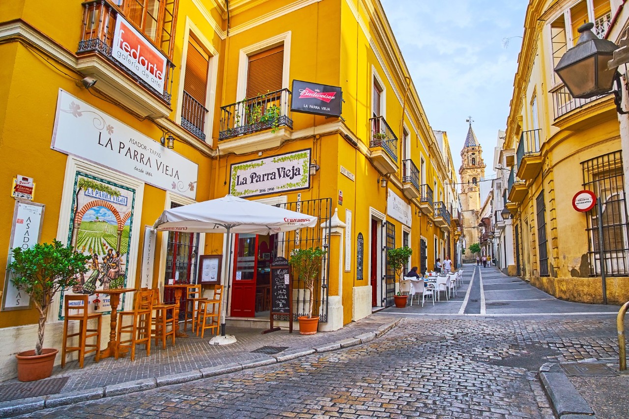 The picture shows a cobbled street with sun-yellow old town houses. On the left-hand side of the street, there are restaurants and bars with tables and chairs outside. A church steeple can be seen in the background. 