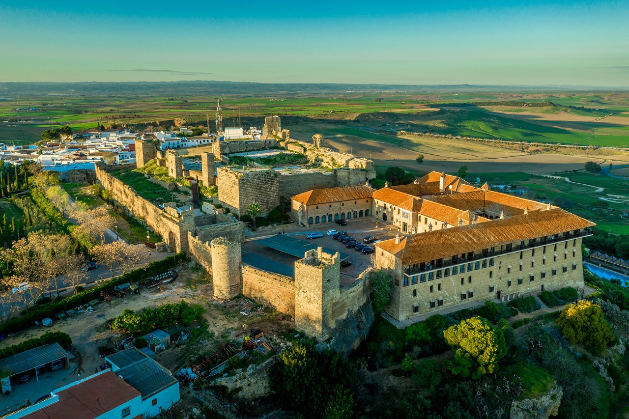 Aerial view of a medieval fortress with towers and walls, adjacent to a large building with red tiled roofs, surrounded by fields and a rural area. 