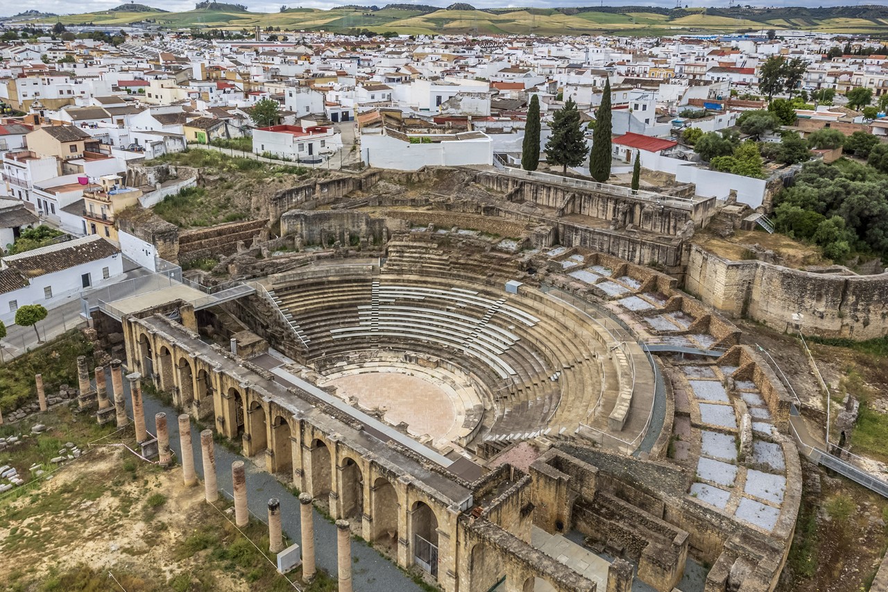 Bird's-eye view of an ancient Roman amphitheatre surrounded by a white village landscape with green hills in the background.