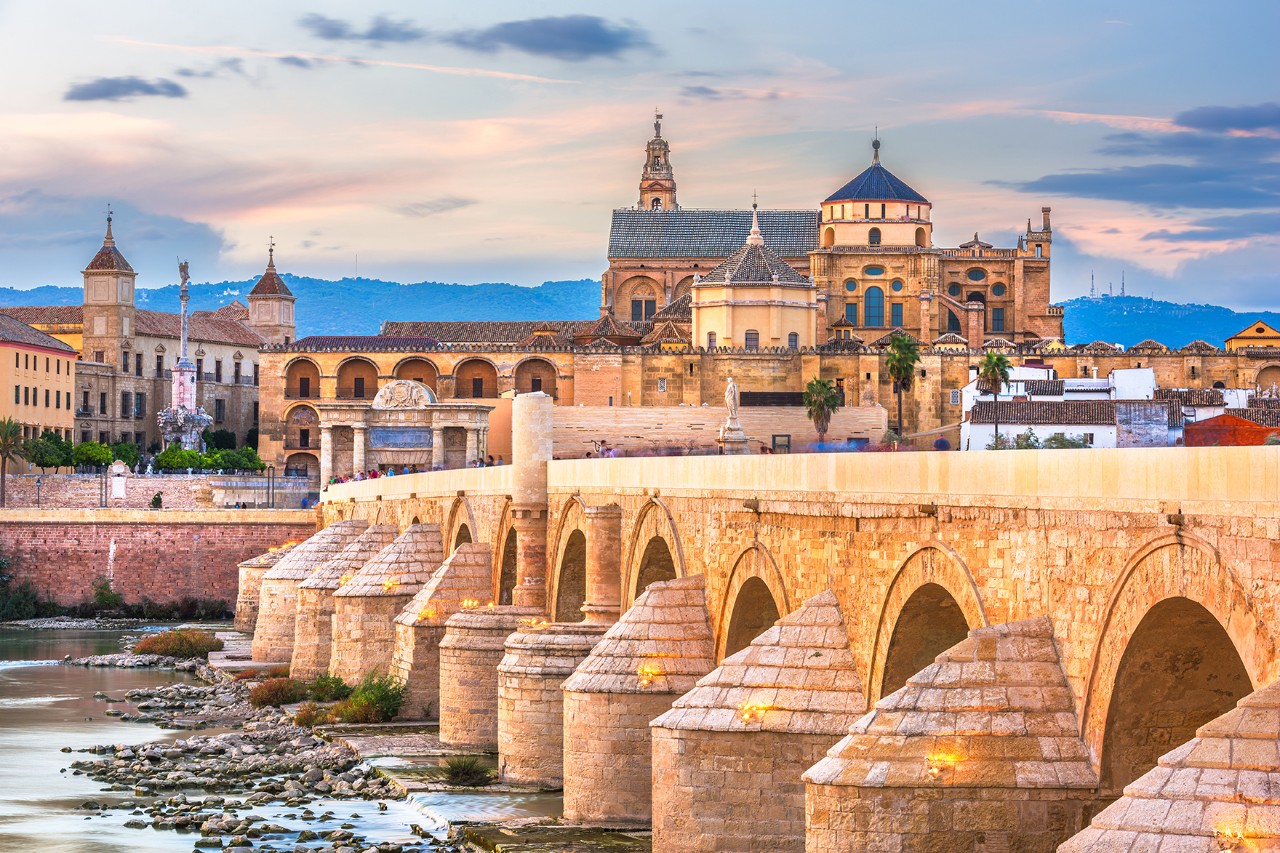 Old stone bridge with round arches over a river, with an imposing cathedral standing on the opposite bank at sunset.