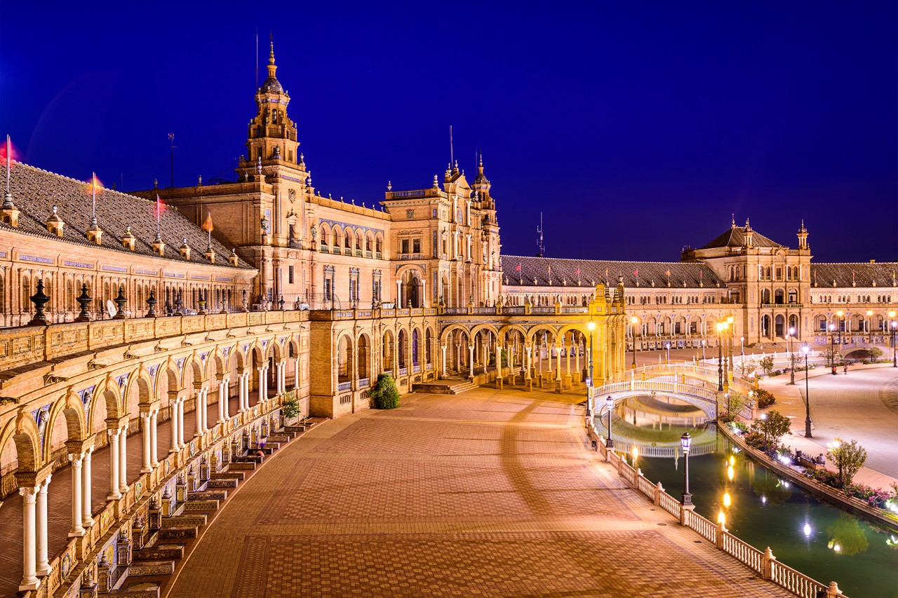 A square illuminated at night with a semicircular building, arcades, bridges and a greenish shimmering water channel.