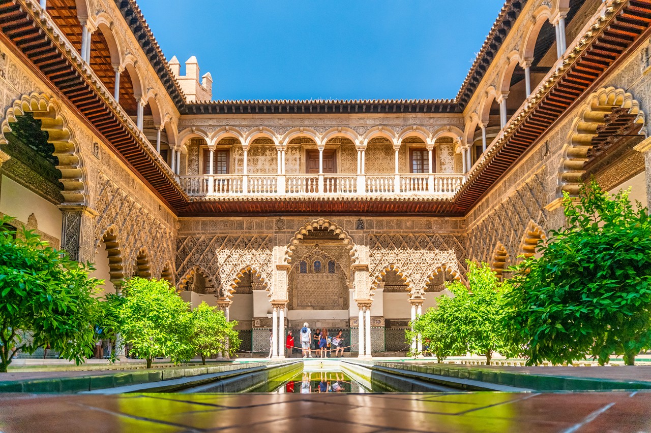 Inner courtyard of a Moorish palace with ornate arches, green bushes and a narrow water basin.
