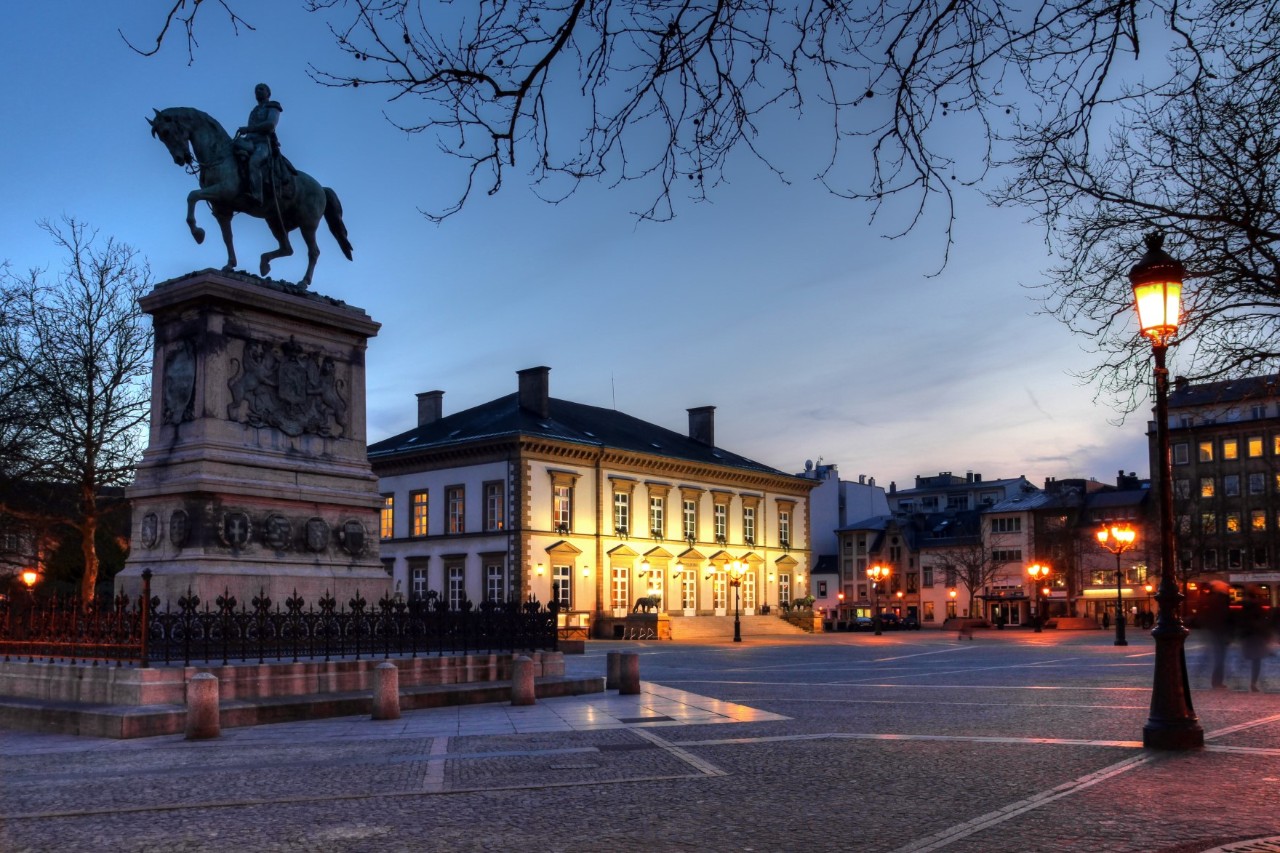 Evening atmosphere at the deserted Place Guillaume II with equestrian statue, street lamps and illuminated buildings © Bogdan Lazar/stock.adobe.com 