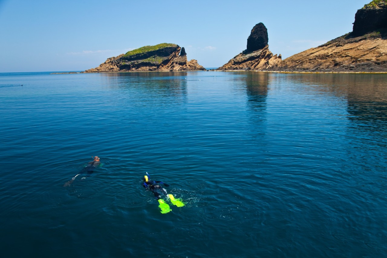 Two people snorkelling on the surface of the sea, barren island mountains in the background 