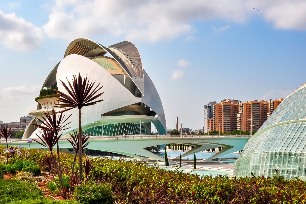 Modern dome-shaped building surrounded by palm trees, green space, a bridge, a glass building and red residential buildings 