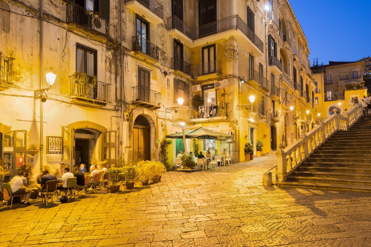 Evening photograph of a cobbled square in the warm light of the street lamps. Several old buildings can be seen on the left of the picture, including street cafés where people are sitting. 