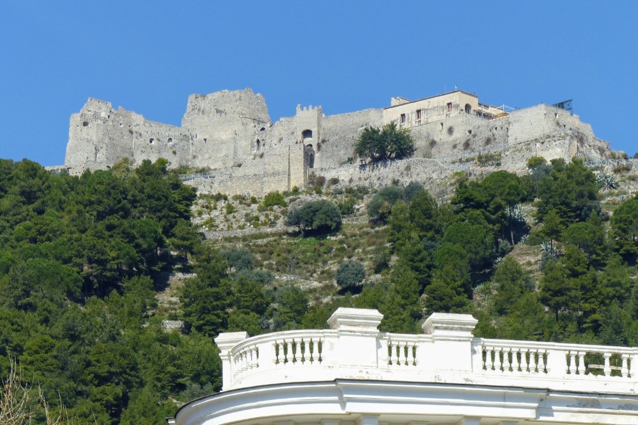 View from below of a white-grey fortress on a densely overgrown hill. A roof terrace of a building surrounded by a white balustrade can be seen in the foreground.