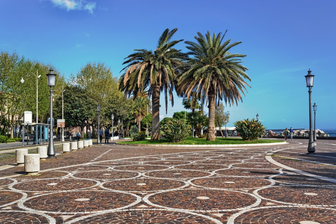 Picture of a square with a terracotta-white patterned mosaic floor. There are historic street lamps to the right and left, and a grass roundabout with several palm trees can be seen in the centre of the picture. 