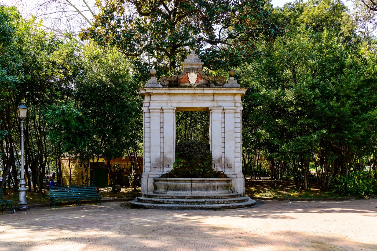 View from an unpaved square to a fountain with a marble superstructure above it. Behind the fountain is a lush park with trees and bushes. 