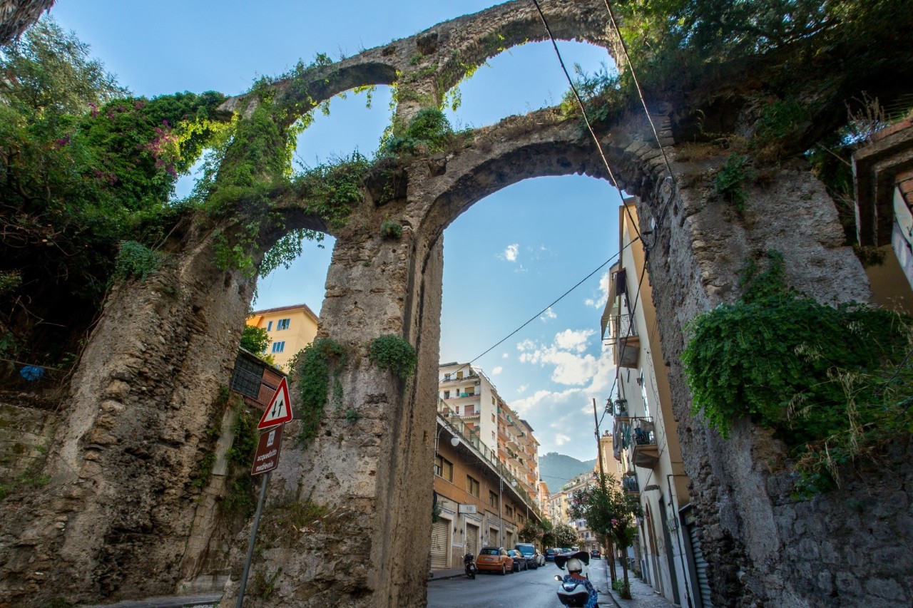 View of very high arcades with double arches in the centre of a city. The obviously historic arcade construction is partly overgrown and a road leads through two high arches.