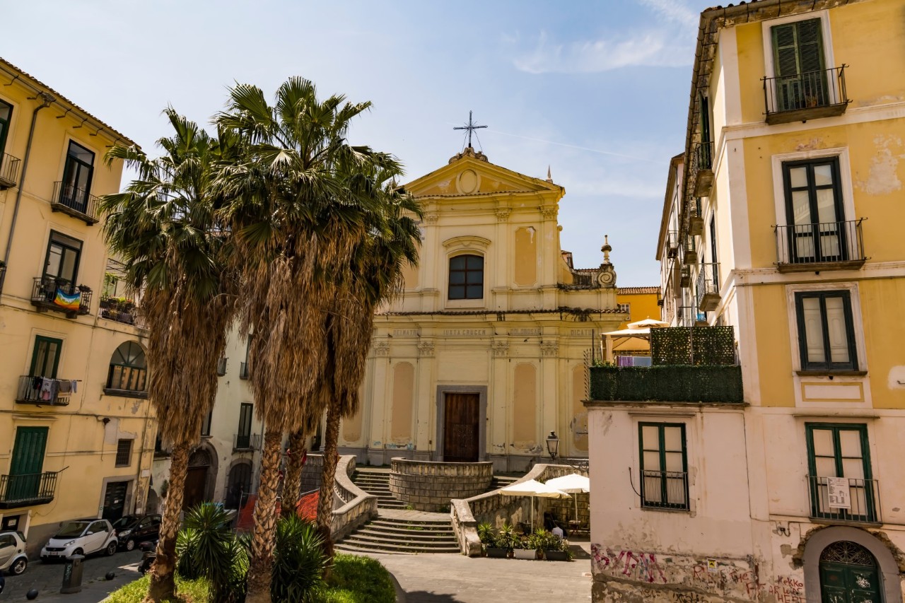 View of the yellow front of a church to which a short, circular staircase leads upwards. There is a cross on the roof. There are palm trees on the square in front of the church and parasols below the steps. 